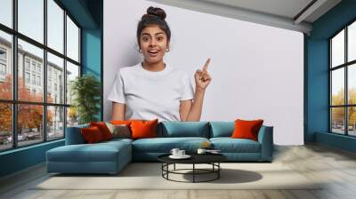 Horizontal shot of pretty surprised cheerful young woman pointing to empty copy space advertises product or tells about awesome offer dressed in casual clothing isolated over white background Wall mural