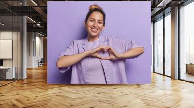 Horizontal shot of pretty young European woman makes heart hand gesture demonstrates love sign smiles tenderly wears shirt and golden earrings poses against purple background. Romance concept Wall mural