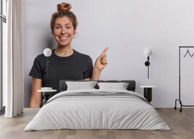 Horizontal shot of pretty young European woman with hair bun smiles pleasantly dressed in casual black tshirt points index finger on empty space isolated over white background. Look at this. Wall mural