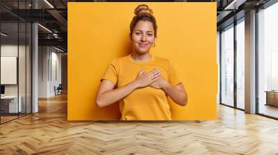 Horizontal shot of pretty young woman presses hands to heart expresses gratitude expressing appreciation and kindness smiles happily dressed in casual t shirt isolated over yellow background Wall mural