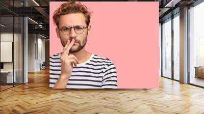 Horizontal shot of serious displeased man frowns face, keeps hand on mouth, has curly hair and stubble, wears striped t shirt, round spectacles, stands against pink background with copy space Wall mural