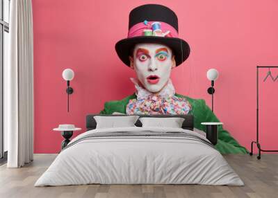 Horizontal shot of stupefied crazy man has image of hatter drinks hot tea poses with cup wears hat and green costume entertains on halloween party. Comic book character holds beverage stands indoor Wall mural