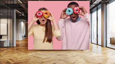 Horizontal shot of surprised dark skinned man covers eyes with delicious sparkling doughnuts, stands near her girlfriend, spend free time together, like eating dessert, isolated over pink background Wall mural