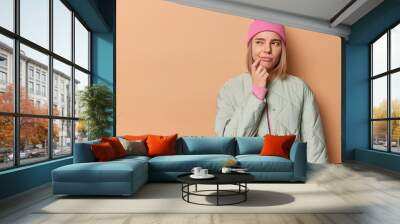 Horizontal shot of thoughtful woman considers something and looks away keeps finger near corner of lips wears pink hat and jacket isolated over brown background copy space on left feels hesitant Wall mural