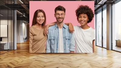 Horizontal shot of three mixed race teenagers spend time together, pose for common photo against pink background. Satisfied guy in eyewear and denim shirt stands between two cheerful women indoor Wall mural