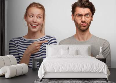 Horizontal shot of two female and male best friends express different emotions, pose together against white blank wall. Cheerful young woman and puzzled unshaven male indicate at each other. Wall mural