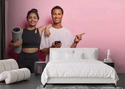 Horizontal shot of two sporty woman and man dressed in sportswear carry foam roller for massaging point index finger on copy space for your advertising content isolated over pink background. Wall mural