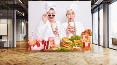 Horizontal shot of two women drink fizzy drink with straws wear domestic comfortable dressing gown cant resist craving for junk food pose near table full of high calorie cakes burgers french fries Wall mural