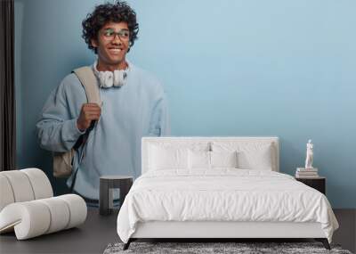 Horizontal studio photo of young delighted smiling Hindu wearing casual blue hoodie and jeans with white bag isolated on left on blue background looking at blank space for your advertisement Wall mural