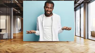 Horizontal view of happy careless guy, spreads hands with clueless facial expression, has no idea, makes choice between two pleasant things, wears white stylish outfit, models over blue background. Wall mural