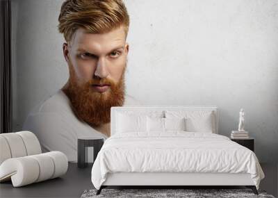 Human face expressions and emotions. Headshot of young model with thick beard posing in studio with angry and unfriendly look. Bearded hipster dressed in white t-shirt with rolled up sleeves frowning Wall mural