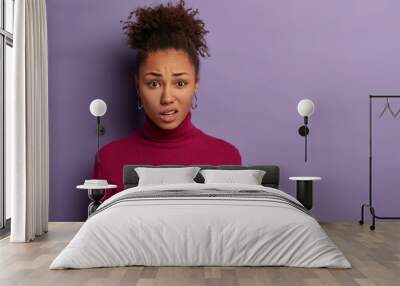 Human facial expressions concept. Displeased Afro woman smirks face and purses lips, reacts on hearing something unpleasant, wears casual jumper, models over purple wall with blank space aside Wall mural