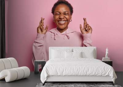 I have to win. Positive dark skinned woman with broad smile and closed eyes raises fingers crossed makes desirable wish waits for good news has white even teeth wears hoodie isolated over pink wall Wall mural