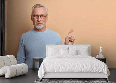 I recommend you to use this copy space. Self confident grey haired European man points at upper right corner shows great place dressed in casual blue jumper isolated over brown studio background Wall mural