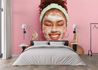 Image of happy ethnic woman raises palms over face, keeps eyes closed, shows white teeth, uses cleansing foam for skin care, gets real pleasure, wears headband and earrings, stands topless alone Wall mural