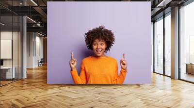 Image of positive dark skinned young lady with Afro hairstyle, points with both index fingers up, dressed in orange jumper, isolated over purple background, has impressed surprised expression Wall mural