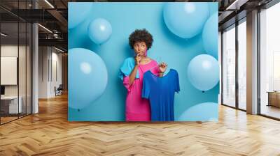 Indecisive thoughtful African American woman chooses outfit for banquet, thinks what to wear, holds dress on hangers, isolated over blue background with big balloons. Women, fashion, clothing Wall mural