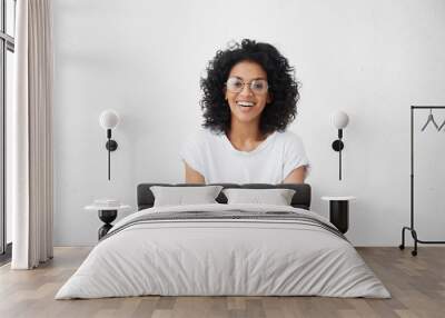 Indoor portrait of beautiful young dark-skinned woman wearing big round eyeglasses and casual white t-shirt smiling joyfully, keeping arms crossed, laughing out loud while watching comedy on TV Wall mural