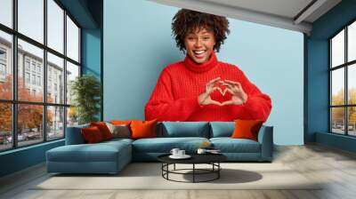 Indoor shot of attractive female model shapes heart symbol with both hands, demonstrates gesture of love to someone, wears red winter clothes, smiles positively, isolated over blue background Wall mural