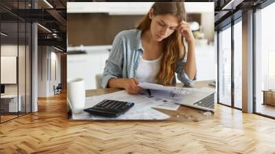 Indoor shot of casually dressed young woman holding papers in her hands, calculating family budget, trying to save some money to buy new bicycle to her son, having stressed and concentrated look Wall mural