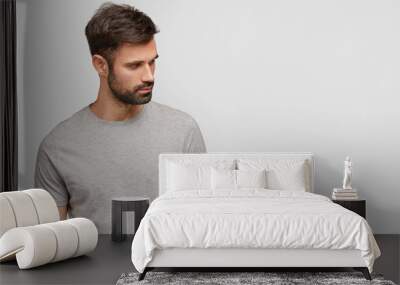 Indoor shot of concentrated handsome unshaven young male foused aside, has muscular body, dressed in casual grey t shirt, stands against white background with blank space for your advertisement Wall mural