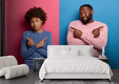 Indoor shot of confused ethnic couple cross hands over chest, point left and right, show directions, cannot make choice, have different opinions towards something, wears blue and pink jumpers Wall mural