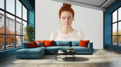 Indoor shot of cute redhead girl looking away, having doubtful and indecisive face expression, pursuing her lips as if forbidden to say anything. Confused young female posing isolated at white wall Wall mural