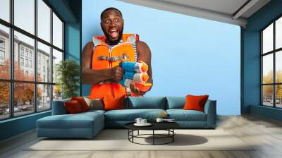 Indoor shot of emotional dark skinned man has fight with someone, plays with water gun, stares into distance with widely opened mouth, enjoys real battle, wears protective lifevest. Summer activity Wall mural