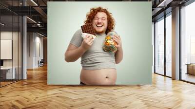 Indoor shot of fat redhead male holding glass of sweets in one hand and bar of chocolate in other, laughing, having sly look, anticipating eating sweets, his belly sticking out of undersized t-shirt Wall mural