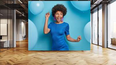 Indoor shot of happy carefree woman dances and has fun dressed in fashionable clothes has festive mood poses against blue background with big inflated balloons around. Time for having party. Wall mural