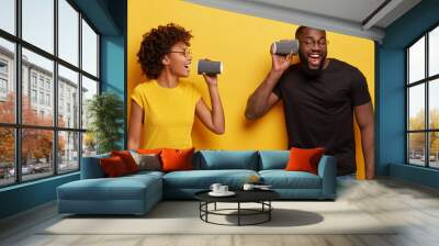 Indoor shot of happy dark skinned woman holds disposable cup near mouth, pretends singing, optimistic guy carries paper container near ear, have fun together after drinking coffee, isolated on yellow Wall mural