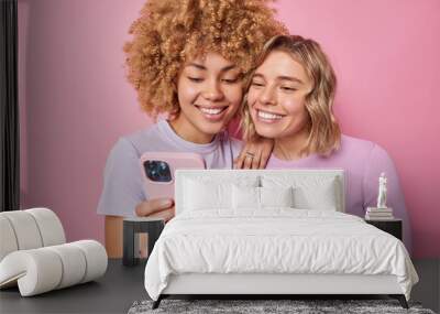 Indoor shot of happy friendly women use cool gadget and application watch video online smile gladfully dressed in casual clothes isolated on pink background enjoy texting and networking communication Wall mural