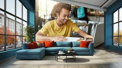 indoor shot of happy student male with curly hair dressed casually sitting in cafeteria working with Wall mural