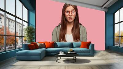 Indoor shot of indignant woman frowns face in displeasure, wears spectacles, shirt, models against pink background, doesnt like something dressed in casual shirt. Facial expressions concept. Wall mural