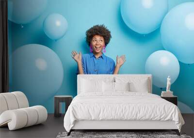 Indoor shot of positive curly haired Afro American woman raises palms dressed in festive dress looks above isolated over blue background with inflated balloons around enjoys birthday celebration Wall mural