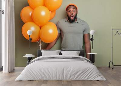 Indoor shot of shocked dark skinned muscular man stares with mouth opened holds bunch of orange inflated balloons dressed in casual clothes reacts on unexpected congratulations. Party attributes Wall mural