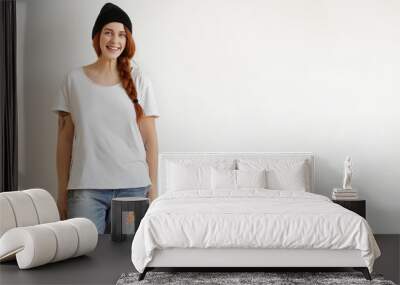 Indoor shot of stylish Caucasian young redhead woman wearing trendy black hat, white t-shirt and ragged jeans standing isolated against blank studio wall with copy space for your advertising content Wall mural