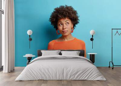 Indoor shot of thoughtful Afro American woman concentrates somewhere pensively has curly hair dreams about something wears casual orange jumper isolated over blue background. Pensive ethnic girl Wall mural