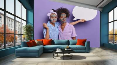 Indoor shot of two thoughtful women apply nourishing clay masks stand closely to each other dressed in pajama undergo beauty treatments at home show blank communication bubble for your text. Wall mural