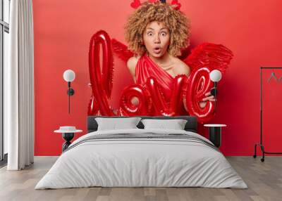 Indoor shot of young African american woman wearing costume of Cupid celebrating St. Valentine's Day organising party surprised having so many guests looking straight at camera with love sign Wall mural