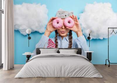 Indoor shot of young birthday girl keeps glazed delicious doughnuts over eyes keeps lips rounded has two pigtails wears blindfold and shirt isolated over blue background. Woman holds tasty donuts Wall mural