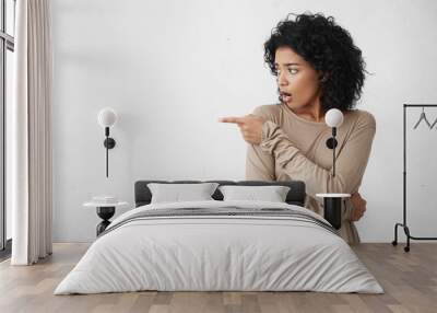 Indoor studio shot of shocked or surprised casually dressed mixed race brunette woman indicating something astonishing on white blank wall, pointing her finger and keeping mouth wide opened Wall mural