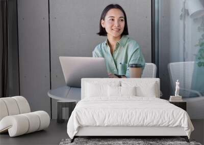 Intelligent female student of university searches information for course work uses modern laptop device looks thoughtfully away poses at small table with notebooks and takeaway coffee checks database Wall mural