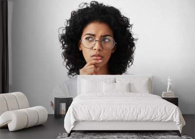 Isolated portrait of stylish young mixed race woman with dark shaggy hair touching her chin and looking sideways with doubtful and sceptical expression, suspecting her boyfriend of lying to her Wall mural