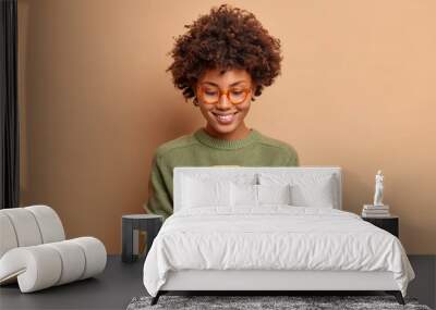 Isolated shot of African American woman uses smartphone application enjoys browsing social media creats news content makes online order wears spectacles and casual jumper poses over beige studio wall Wall mural