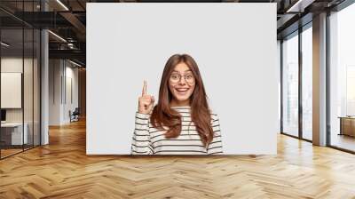 Isolated shot of cheerful Caucasian woman with straight hair, shows free space, points with index finger for your advertising content, wears casual striped sweater, isolated over white background Wall mural