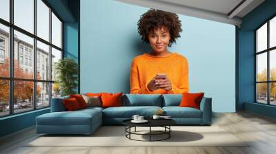 Isolated shot of positive smiling woman holds modern cellular, enjoys wireless internet, checks bank account or sends message in social networks, wears orange clothes, models over blue background Wall mural