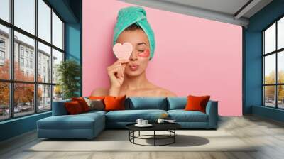 Isolated shot of pretty female model has eyes closed, lips rounded, stands with bare shoulders, enjoys beauty treatments, holds cosmetic sponge over eye, cares about face wears hydrogel patches, towel Wall mural