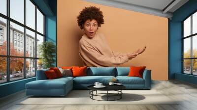 Isolated shot of surprised young Afro American woman pretends catching something big, keeps palms, holds invisible object from under, dressed in casual sweatshirt, isolated on beige background. Wall mural