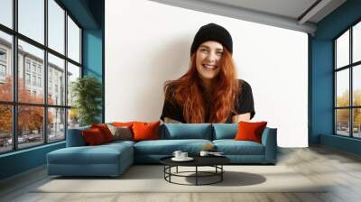 Isolated studio portrait of fashionable female model with gorgeous long ginger hair looking and smiling at camera, standing against white wall. Pretty girl in stylish clothes posing indoors Wall mural